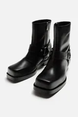 Bota negra de cuero con caña corta, horma cuadrada, suela a tono con ligero tacón y arnés decorativo a la altura del tobillo con detalles metálicos.