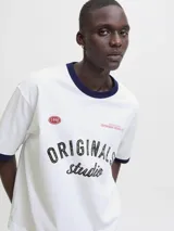 Remera blanca de manga corta y cuello redondo, con fit oversize y largo cropped. Tiene ribetes azul marino en el cuello y las mangas. Estampado frontal con la palabra "ORIGINALS" en negro y "studio" en cursiva, junto a detalles en rojo como "1990" y "COPENHAGEN CREATIVE DEPT.".