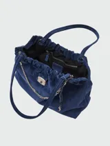 Bolso tote de gamuza color azul marino, con frunce ajustable en la parte superior y asas de hombro. Presenta herrajes metálicos plateados, incluyendo un dije de cadena decorativo y un cierre de solapa con logo grabado. El interior es forrado en tela negra y cuenta con un bolsillo con cierre.