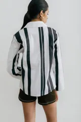 Camisa blanca con rayas verticales negras, de manga larga y corte recto. Tiene cuello clásico, cierre con botones y bolsillo en el pecho.