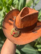 Sombrero de ala ancha tejido en hilo de coco y sintético, color marrón claro. La copa está cubierta con encaje naranja quemado con patrón floral. Tiene una cinta de cuero marrón oscuro y un aplique decorativo de hoja de gamuza con incrustaciones de pedrería.