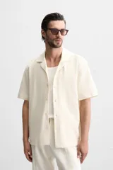 Camisa color crudo de corte regular fit, confeccionada en tejido con textura. Tiene cuello bowling, manga corta, bajo con aberturas laterales y cierre frontal con botones.