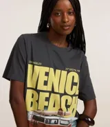 Remera gris con estampa de la frase "Venice Beach" en amarillo y una imagen de una camioneta con tablas de surf.