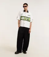 Camisa blanca oversized de tricolina con cuello negro contrastante, mangas cortas y estampado con la palabra "Hustle" en verde y negro.