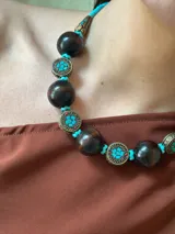 Collar corto con cuentas grandes de madera color marrón oscuro, separadores de metal con incrustaciones de turquesas y cuentas pequeñas turquesas. Cierre con cadena de acero quirúrgico y ganchito para regular el largo.
