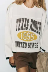 Sudadera blanca de algodón con cuello redondo y mangas largas. Estampado frontal con texto "Texas Rodeo 1995 United States" en color marrón.