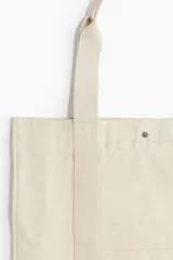 Bolso shopper de lona color beige con doble asa de hombro y cierre de botón a presión. Tiene un compartimento exterior y un compartimento interior pequeño, ambos abiertos.