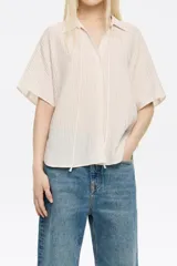 Camisa color beige estilo capa confeccionada en viscosa con textura. Cuenta con cuello camisero con lazo, manga corta murciélago y cierre frontal con botones.