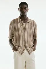 Camisa de manga corta con cuello inglés, botones adelante y bajo recto. Tejido de punto pointelle con estilo crochet y diseño a rayas verticales en tonos beige, marrón y blanco.