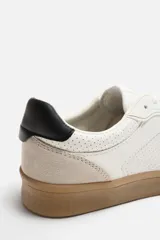 Zapatillas deportivas de estilo retro, color blanco con detalles en beige y negro. Capellada de cuero sintético con perforaciones laterales y puntera de gamuza. Suela de goma color caramelo.