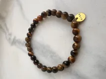 Pulsera elástica de piedras naturales color marrón con dije circular dorado con la palabra "AIRE" grabada.