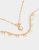Collar corto doble, con cadena dorada de eslabones y cadena con dijes de moños. Incluye cierre marinero con dije de corazón de perla.