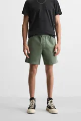 Bermuda color verde militar de corte relajado, confeccionada en tejido de algodón. Tiene cintura elástica ajustable con cordón interior, bolsillos frontales y bolsillo trasero de parche.