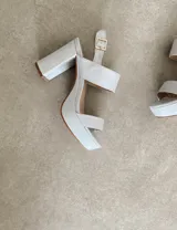 Sandalias de tacón alto color blanco, con plataforma y tira ancha sobre los dedos. El tobillo se sujeta con una tira fina con hebilla dorada. El tacón es ancho y cuadrado.