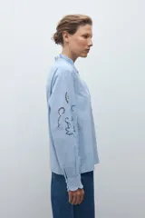 Camisa de algodón celeste claro, de manga larga, con cuello mao con volados y bordados calados florales en el frente y las mangas.