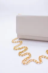 Cartera tipo sobre color beige con textura granulada y correa de cadena dorada.