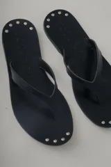 Sandalia plana tipo flip flop de cuero negro, con diseño minimalista y tira central de cuero. Presenta detalles de tachas metálicas en la base.