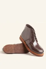 Bota de cuero vacuno con acabado charol, color marrón oscuro. Presenta un diseño de caña corta con cordones frontales y costuras decorativas en la puntera. La suela es de goma tipo crepe.