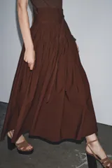 Vestido midi marrón con cuello barco y manga sisa. Presenta un corte a la altura de la cintura con detalle de trabilla y lazo del mismo tejido. Cierre lateral con cremallera oculta.