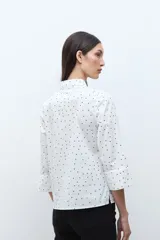 Blusa blanca con estampado de pequeños lunares negros, cuello camisero y botones frontales oscuros. Mangas tres cuartos con ligero vuelo en el puño.