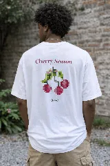 Remera blanca de algodón premium con estampado en la espalda que dice "Cherry Season" y un diseño de cerezas en estilo pixel art, con un pequeño logo ovalado con la palabra "tuya" debajo.