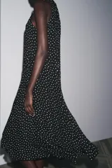 Vestido midi negro con estampado de lunares blancos, cuello halter con pañuelo integrado, sin mangas y bajo asimétrico. Cierre con abertura y botón en la nuca.