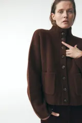 Chaqueta de punto marrón con cuello alto y manga larga. Tiene cierre frontal con botones y bolsillos de parche en el delantero.