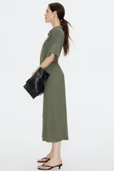 Vestido midi color verde oliva con cuello camisero, botonadura frontal y mangas raglán cortas. Tiene elástico fino en la parte posterior de la cintura, frunces adelante y atrás, y cinturón extraíble para atar.