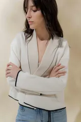 Chaqueta de punto color crudo con solapa ancha acanalada terminada con cadenetas en contraste negro. Tiene calce relajado, hombros caídos, largo a la cadera y manga larga.
