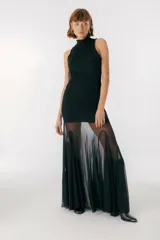 Vestido largo color beige, con cuello alto y sin mangas. La parte superior es ajustada al cuerpo y la falda es amplia y fluida, con paneles traslúcidos.