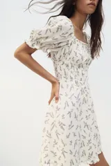 Vestido corto blanco con estampado floral en tonos lavanda, escote cuadrado con volantes, mangas cortas abullonadas con volantes y cuerpo fruncido.