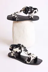 Sandalias planas con tiras de cuero con estampado de vaca en blanco y negro, y hebilla lateral.