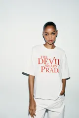 Camiseta blanca de algodón con cuello redondo, mangas cortas y hombros caídos. Estampado frontal con el título de la película "El Diablo Viste de Prada" en letras rojas.