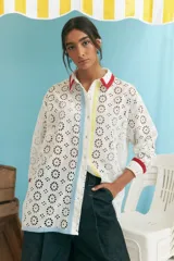 Camisa blanca oversize de algodón con bordado calado tipo broderie, cuello camisero con ribete rojo, cierre frontal con botones y mangas largas con puños con ribete rojo. Presenta una franja vertical con ribete celeste y otra con ribete amarillo.