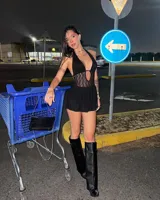 Vestido corto negro con escote halter profundo y detalle de encaje o red en el torso. La falda es corta y con textura. Se combina con botas altas negras de cuero sintético.