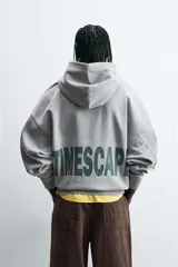 Sudadera gris con capucha ajustable, corte boxy fit, manga larga, bolsillo tipo canguro y estampado en la espalda.