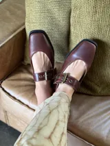 Zapatos de cuero color marrón con punta cuadrada, tira sobre el empeine con hebilla dorada y suela plana.