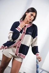 Cardigan tejido a rayas horizontales rojas y crema, con cuello redondo, cierre frontal con botones marrones y mangas largas abullonadas. Presenta ribetes a rayas en cuello, puños y ruedo. Tiene bordado el logo de la marca en el pecho.