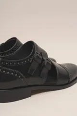 Zapato bajo de cuero negro, estilo monk strap con dos hebillas ajustables sobre el empeine. Presenta detalles de brogue (perforaciones) en la puntera y alrededor del borde superior. Tiene un taco bajo y ancho.