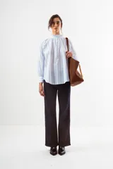 Blusa blanca con finas rayas verticales azules, cuello alto con volado y cierre frontal de botones. Mangas largas abullonadas con puños ajustados.