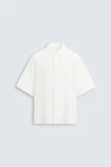 Camisa de hombre de manga corta, cuello solapa y corte relaxed fit. Tejido blanco con textura sutilmente arrugada o plisada. Cierre frontal de botonadura y bajo con aberturas laterales.