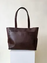 Cartera tipo tote color beige, hecha en cuero graneado con textura uniforme y suave al tacto. Tiene doble asa de hombro, un bolsillo frontal con broche imantado, un bolsillo trasero con cierre metálico y cierre principal también metálico. El interior está forrado y cuenta con tres bolsillos: uno con cierre, uno tipo plaqué para celular y un bolsillo grande tipo sobre con solapa y botón.