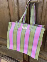 Bolso tipo tote bag con rayas verticales en tonos rosa, celeste y dorado. Tiene asas cortas rosas y etiqueta con la marca "AMÉN".