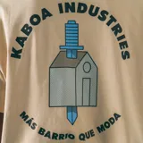 Remera manga corta color crudo, corte oversize, con estampa en la espalda que incluye el logo de la marca Kaboa Industries y la frase "Más barrio que moda".