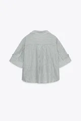 Camisa a rayas verticales grises y blancas, confeccionada en hilatura con lino, de cuello solapa y escote pico. Manga corta con trabilla y botón. Bajo asimétrico. Cierre frontal con botón.