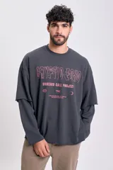 Sudadera de manga larga color gris oscuro con efecto desgastado y dobladillo sin rematar en el bajo. Presenta un diseño de doble manga, donde la manga interior es visible en el puño. Estampado frontal en color rosado con texto temático.