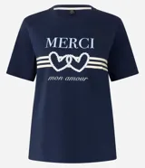 Remera azul marino de algodón con cuello redondo y mangas cortas. Estampa frontal con la palabra "Merci" en color crema, dos corazones entrelazados con detalles de perlas y la frase "mon amour" en cursiva.