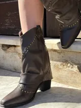 Bota corta de cuero sintético color marrón, con taco cuadrado y punta fina. Presenta una correa con tachas pequeñas y una argolla metálica decorativa alrededor del tobillo. Cuenta con cierre lateral.