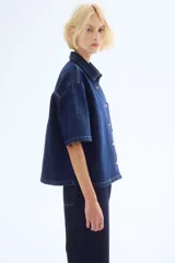 Camisa azul de denim, corte amplio, con cuello clásico, abotonadura frontal, manga corta, bolsillo de parche en el pecho y canesú en la espalda.