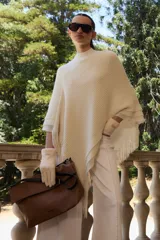 Capa tejida color crudo con cuello alto y flecos en el borde inferior.